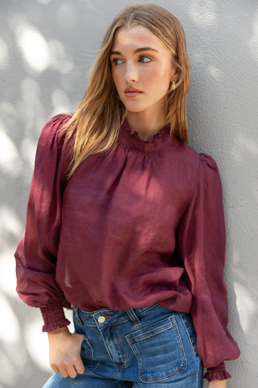 Woman wearing a burgundy blouse and blue jeans against a gray wall.