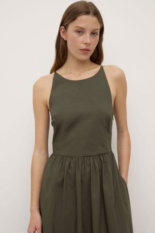 A woman wearing a sleeveless khaki green midi dress with a high neckline, fitted bodice, and gathered waist. The dress has side pockets and a relaxed A-line silhouette, styled simply with black sandals.’