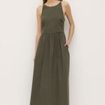 A woman wearing a sleeveless khaki green midi dress with a high neckline, fitted bodice, and gathered waist. The dress has side pockets and a relaxed A-line silhouette, styled simply with black sandals.’