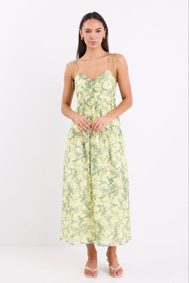 Woman wearing a yellow and green floral print maxi dress with adjustable tie straps, shirred back, and tiered skirt, standing against a white background.