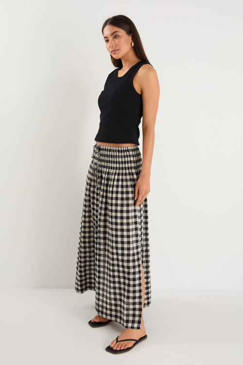 Woman wearing a black gingham maxi skirt with shirred waist, tiered design, and side split, styled with light flat shoes.
