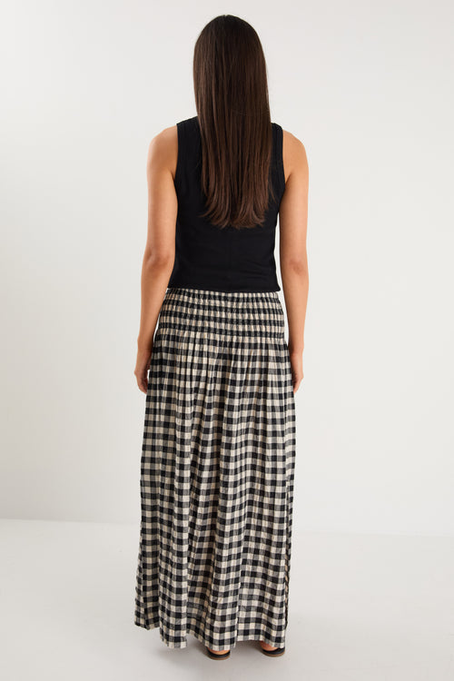 Woman wearing a black gingham maxi skirt with shirred waist, tiered design, and side split, styled with light flat shoes.