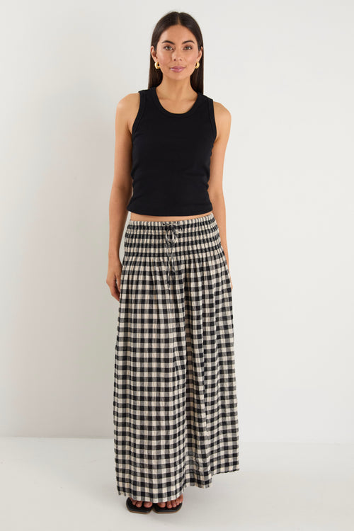 Woman wearing a black gingham maxi skirt with shirred waist, tiered design, and side split, styled with light flat shoes.