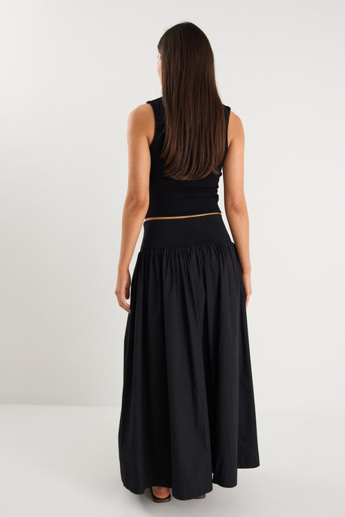 A model wears a black maxi skirt with a ribbed waistband and pleated design, styled with a black sleeveless top and sandals.