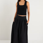A model wears a black maxi skirt with a ribbed waistband and pleated design, styled with a black sleeveless top and sandals.
