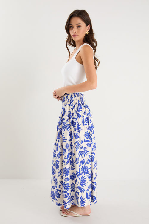 A model wears a long white skirt with a bold blue floral print, elastic waistband, and relaxed fit, paired with a white tank top and neutral sandals.