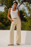 Woman wearing a white tank top and beige pants standing outdoors with greenery in the background