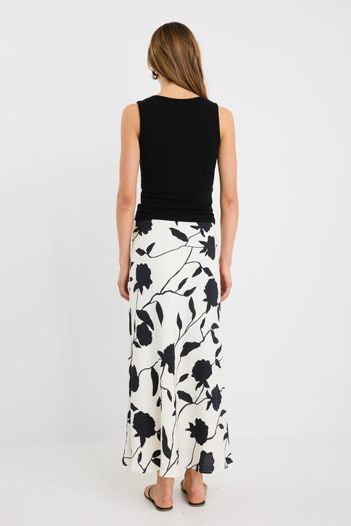 Woman wearing an ivory bias-cut midi skirt with black floral print, elastic waist, and pull-on fit, styled with black sandals and a tucked-in black top.