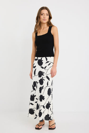 Woman wearing an ivory bias-cut midi skirt with black floral print, elastic waist, and pull-on fit, styled with black sandals and a tucked-in black top.