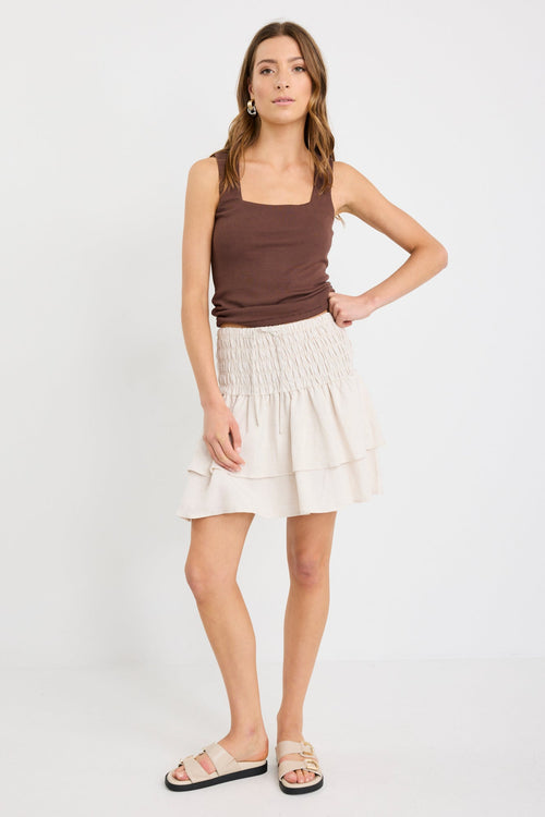 Woman wearing a natural tiered linen-blend mini skirt with a shirred waistband and layered hem, styled with a chocolate tank.