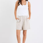 Woman wearing natural linen high-rise tailored shorts with subtle pleats and side pockets, paired with a white tank top and sandals, standing against a white background.