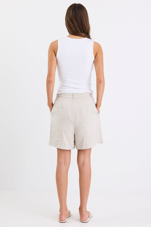 Woman wearing natural linen high-rise tailored shorts with subtle pleats and side pockets, paired with a white tank top and sandals, standing against a white background.