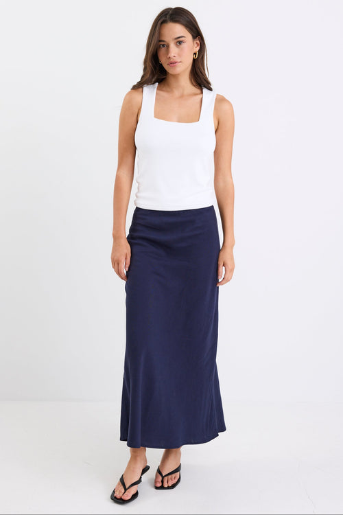 Woman wearing a navy linen bias-cut midi skirt with an elastic waistband, paired with a white tank top and black heeled sandals, standing against a white background.
