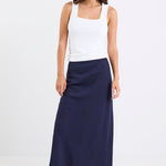 Woman wearing a navy linen bias-cut midi skirt with an elastic waistband, paired with a white tank top and black heeled sandals, standing against a white background.