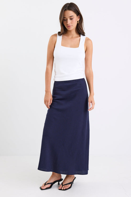 Woman wearing a navy linen bias-cut midi skirt with an elastic waistband, paired with a white tank top and black heeled sandals, standing against a white background.