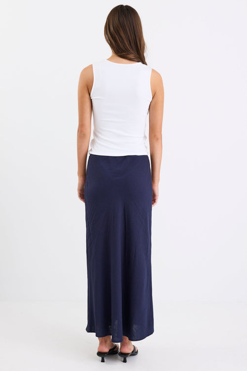 Woman wearing a navy linen bias-cut midi skirt with an elastic waistband, paired with a white tank top and black heeled sandals, standing against a white background.