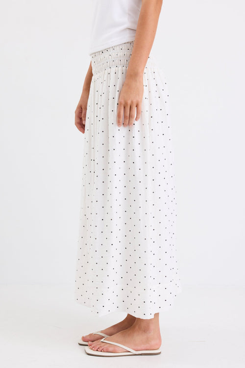 Woman wearing a cream maxi skirt with black polka dots and a shirred elastic waistband, paired with a white tank top and sandals, standing against a white background.