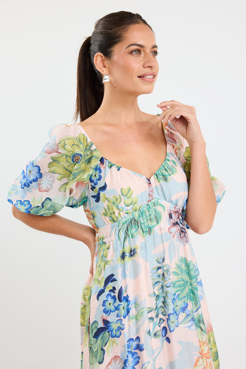 woman wearing a flowing floral puff sleeve dress with elastic waist and pastel blue and green print, styled with tan sandals.