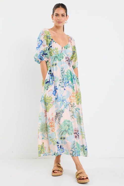 woman wearing a flowing floral puff sleeve dress with elastic waist and pastel blue and green print, styled with tan sandals.