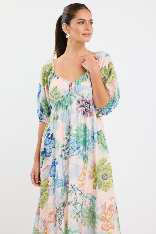 woman wearing a flowing floral puff sleeve dress with elastic waist and pastel blue and green print, styled with tan sandals.