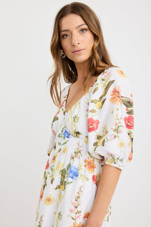 Woman wearing a cream midi dress with a sweetheart neckline, puff sleeves, elastic waist, and garden floral print in red, yellow, and blue tones.