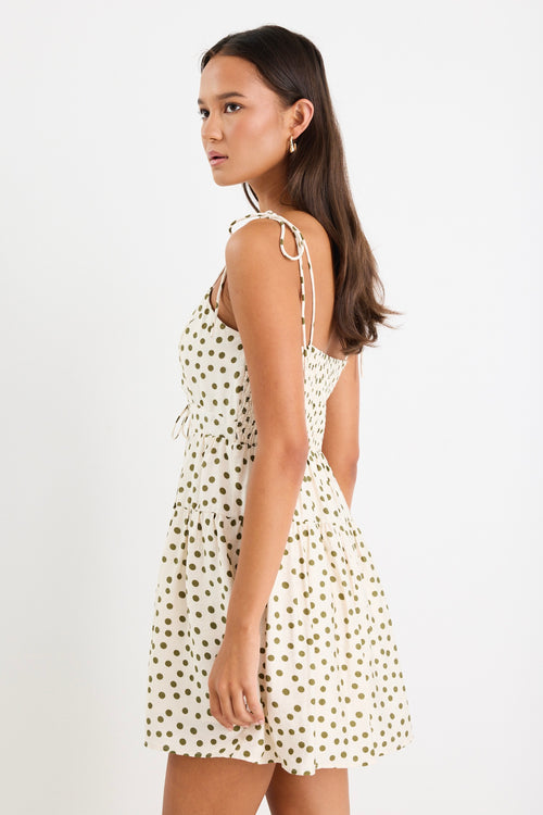 Woman wearing a cream mini dress with khaki polka dots, tiered skirt and tie-up straps, styled with gold jewellery.