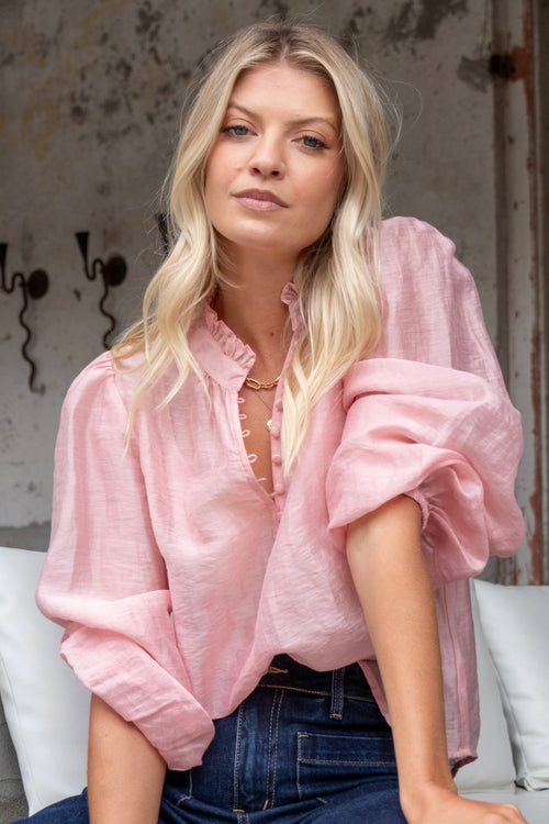 Woman wearing a soft pink blouse with covered buttons and a ruffled collar, paired with dark high-rise jeans.