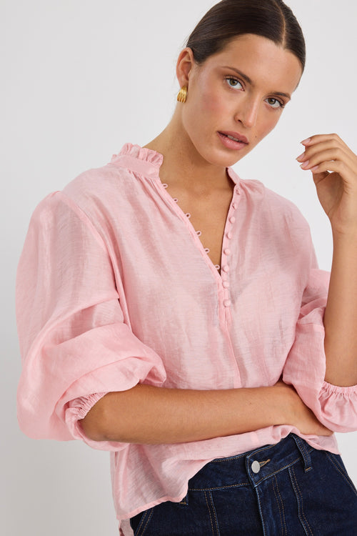 Woman wearing a soft pink blouse with covered buttons and a ruffled collar, paired with dark high-rise jeans.