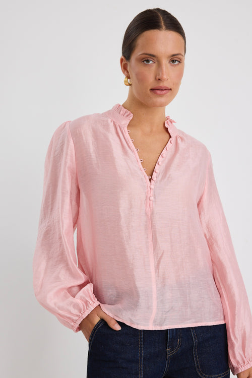 Woman wearing a soft pink blouse with covered buttons and a ruffled collar, paired with dark high-rise jeans.