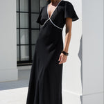 A woman wearing a black maxi dress with white contrast piping stands outdoors in front of a modern white building with columns.