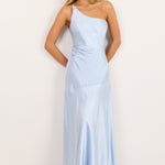 A woman modeling a light blue one-shoulder satin maxi dress against a plain off-white studio background.
