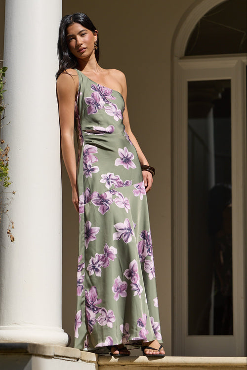 A woman stands on a porch wearing a sleeveless olive green maxi dress with a purple floral print. The dress features a draped halter-style bodice and a flowing skirt.