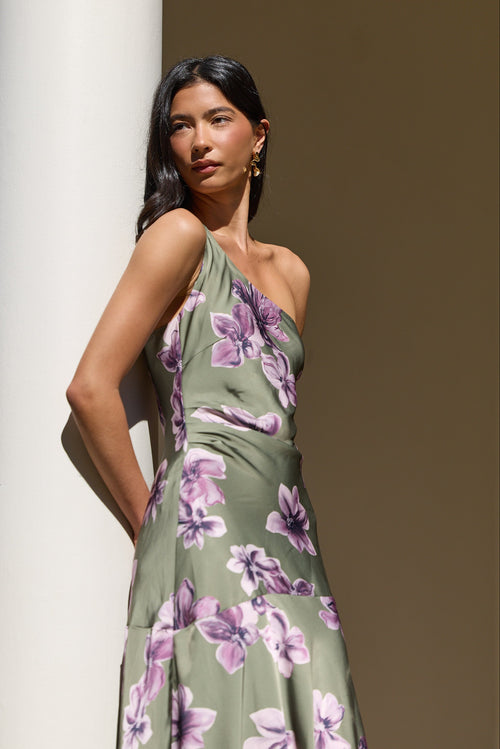 A woman wearing a one-shoulder olive green dress with a purple floral print, posing next to a white column in warm, natural light.