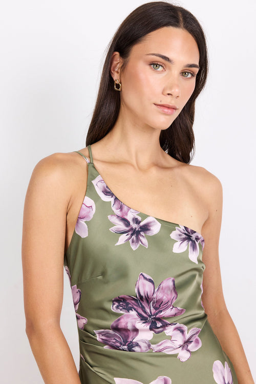 A woman wearing a one-shoulder olive green satin slip dress with a purple floral print, photographed against a plain light gray studio background.