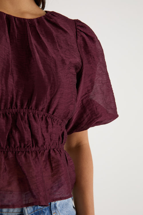 A model wears a burgundy puff sleeve top with a gathered waist, styled with light blue high-waisted jeans.