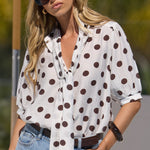 A woman wearing a white short-sleeve button-up blouse with dark brown polka dots, styled with light-wash blue denim shorts, a brown belt, and dark sunglasses.