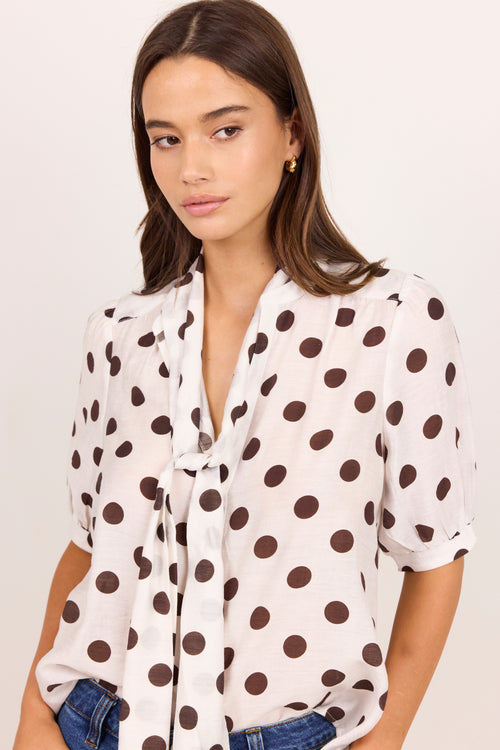 A woman wearing a white short-sleeve blouse with large dark brown polka dots and a tie-neck detail, paired with blue jeans against a plain light background.