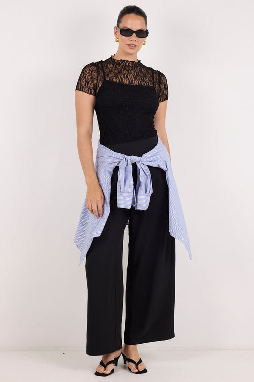 A model stands against a plain white studio background wearing a black short-sleeve top with sheer lace sleeves, black wide-leg pants, and a light blue shirt tied around the waist.