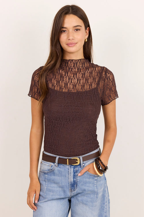 A woman modeling a short-sleeve brown lace and textured knit top with a mock neck, paired with light blue jeans and a brown belt, against a plain off-white background.