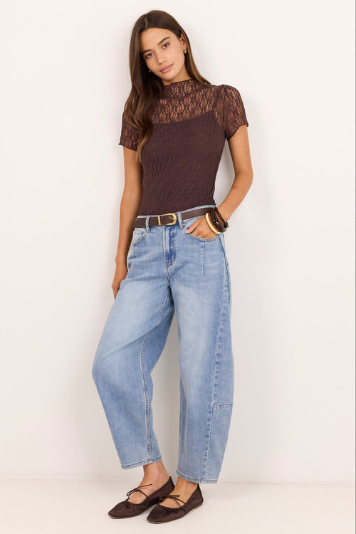 A woman modeling a dark brown short-sleeve top with a sheer lace upper chest and hem, paired with wide-leg light blue jeans and dark brown flat shoes, standing against a plain off-white studio background.