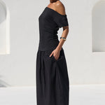 A woman wearing a black off-the-shoulder maxi dress stands on a white tiled poolside patio with a modern white stucco building in the background.