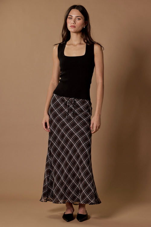 A woman stands against a warm tan studio backdrop wearing a black sleeveless top and a long black skirt with a white plaid pattern.