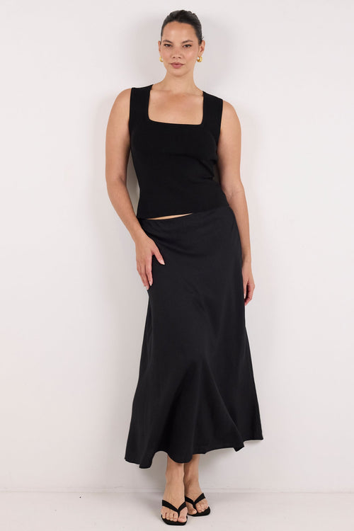 A woman modeling a black sleeveless crop top and matching black maxi skirt against a plain white studio background.