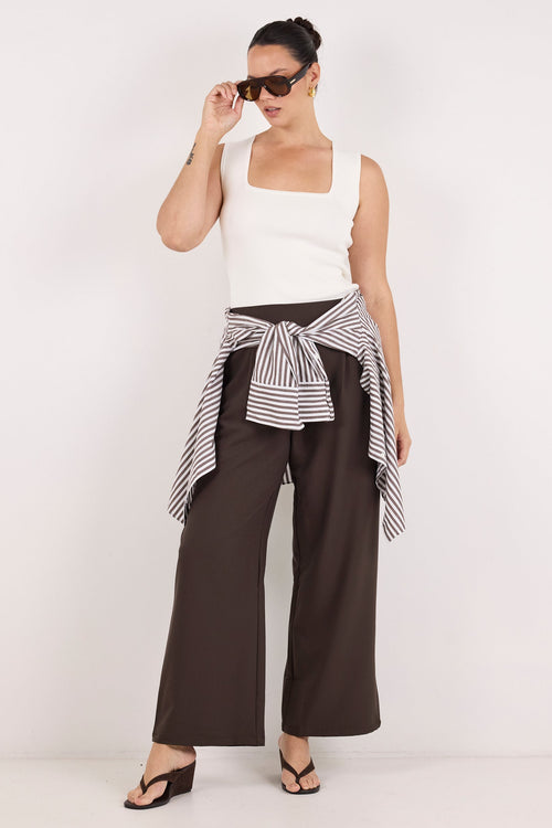 A woman modeling a white sleeveless top, dark brown wide-leg pants, and a black-and-white striped shirt tied around her waist, styled with dark sunglasses and black sandals against a plain white studio background.