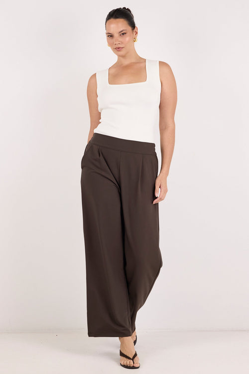 A woman modeling a pair of dark brown wide-leg pants with a white sleeveless top, standing against a plain white studio background.