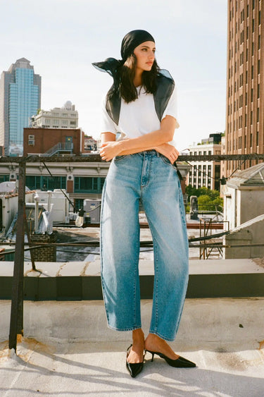 Woman wearing high-rise barrel jeans in a light blue wash, styled with a white tee, heels and a headscarf, standing on a city rooftop.
