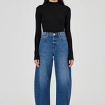 A model stands against a light gray studio background wearing a black long-sleeve mock-neck top, high-waisted wide-leg blue jeans, and black ankle boots.