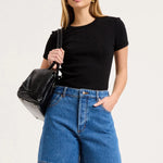 A woman modeling a black short-sleeve crewneck top and blue denim shorts, holding a black shoulder bag against a plain light gray studio background.