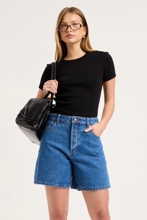 A woman modeling a black short-sleeve crewneck top and blue denim shorts, holding a black shoulder bag against a plain light gray studio background.