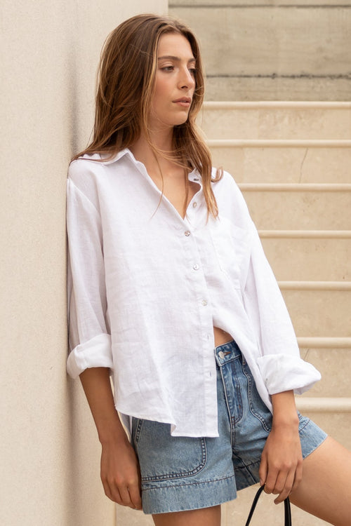 Woman in white shirt and denim shorts standing on stone steps holding a beige bag.
